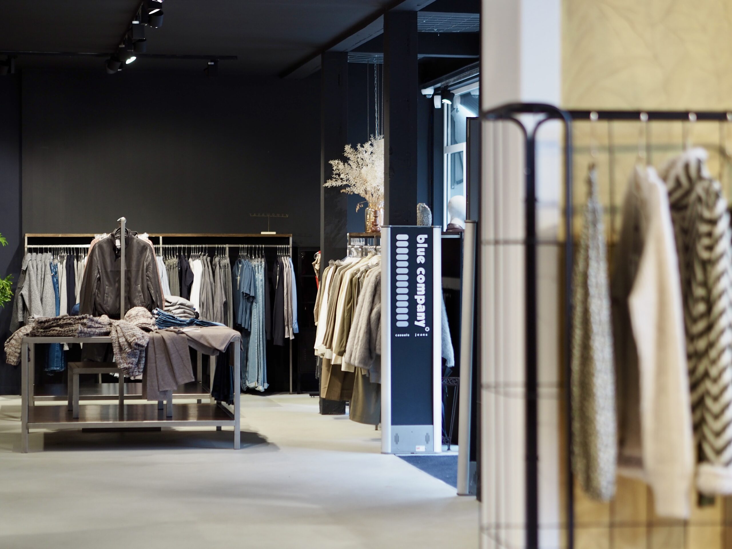 Close‑up van een kledingrek met pastel- en denimitems naast een houten kist met droogbloemen en een foto van een model; geurmarketing versterkt het huiselijke gevoel.